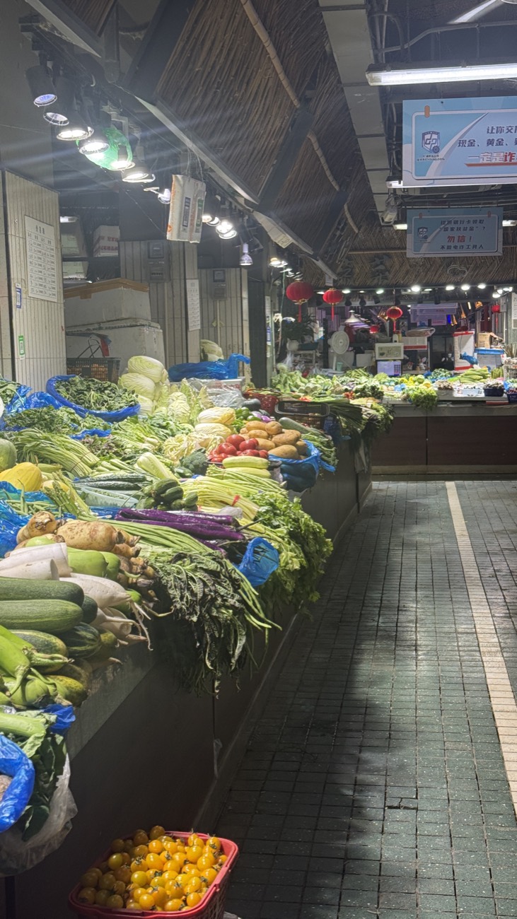 A long produce aisle inside Mengxi Market — stalls of fresh vegetables and fruit running the length of the hall, locals shopping in the morning