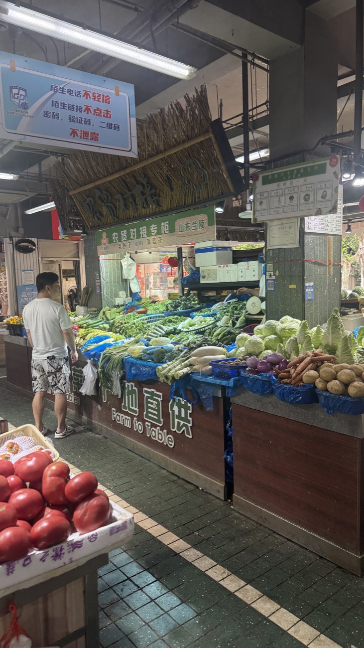 A local shopper at a Mengxi Market produce stall under a 'Farm to Table' sign — crates of tomatoes, leafy greens and root vegetables