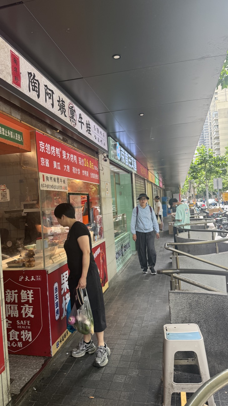 The street-food row just outside Mengxi Market — small shopfronts selling shaobing and grilled snacks, a local carrying a bag of groceries
