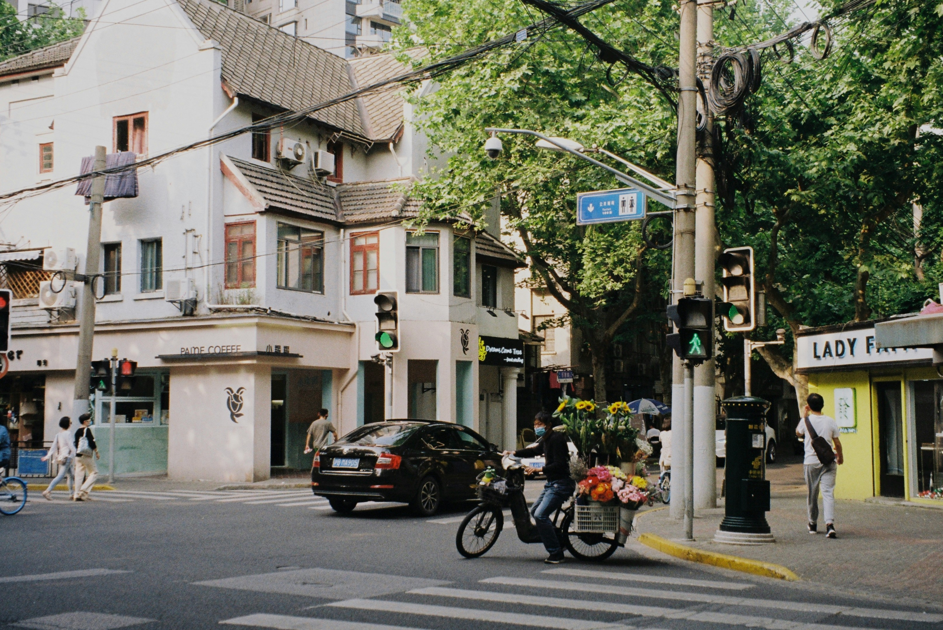 A street corner with PAITIE COFFEE and a flower-cart e-bike at a crosswalk, plane-tree shade overhead — the pivot point between Wukang and Anfu