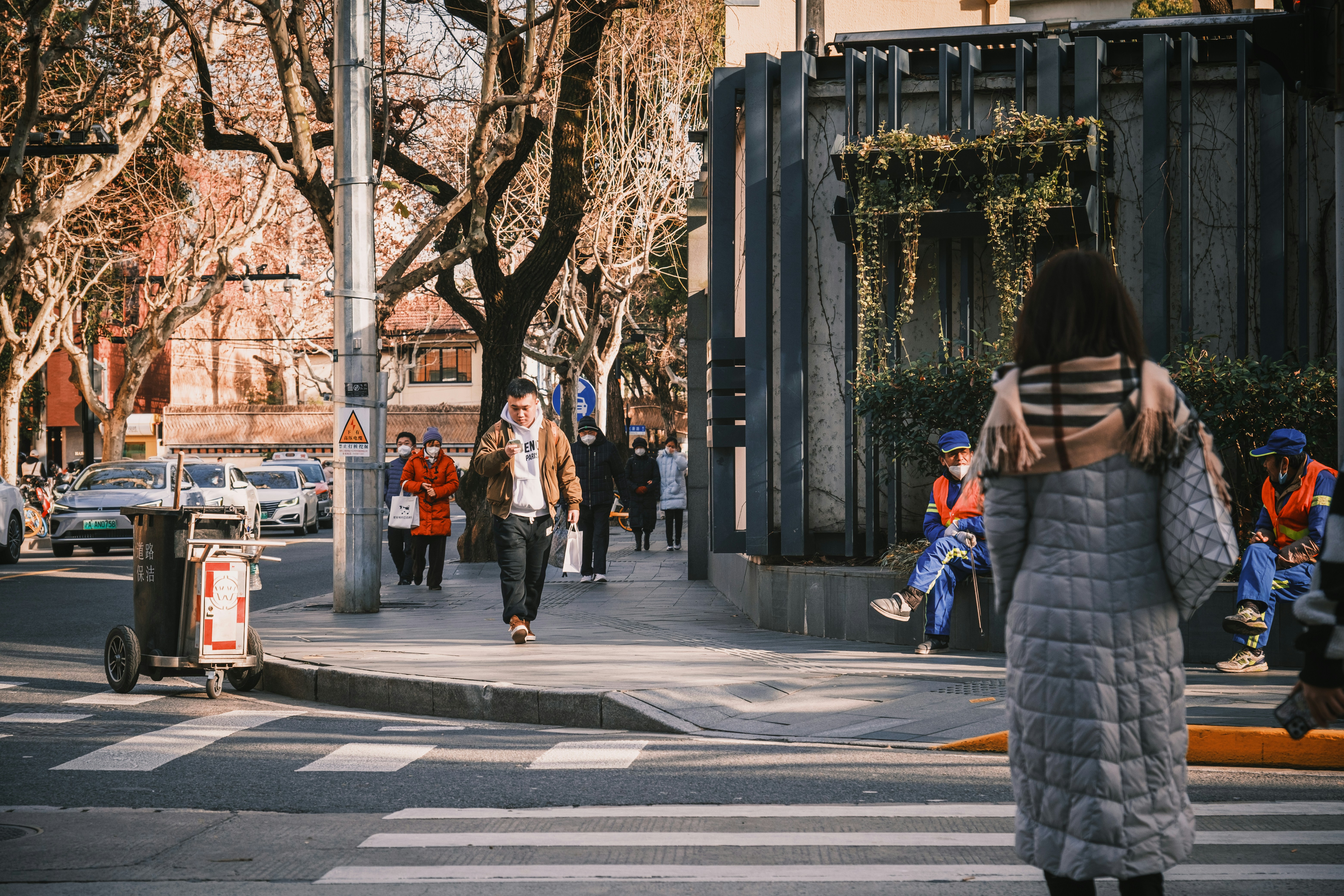 Weekday afternoon scene on Wukang Road — woman in a long coat crossing the street, bare plane trees, sanitation workers resting
